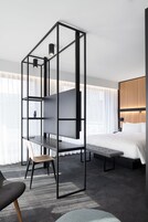 A modern hotel room with a large bed, a desk with a chair, a bookshelf, and a view of the city through large windows.
