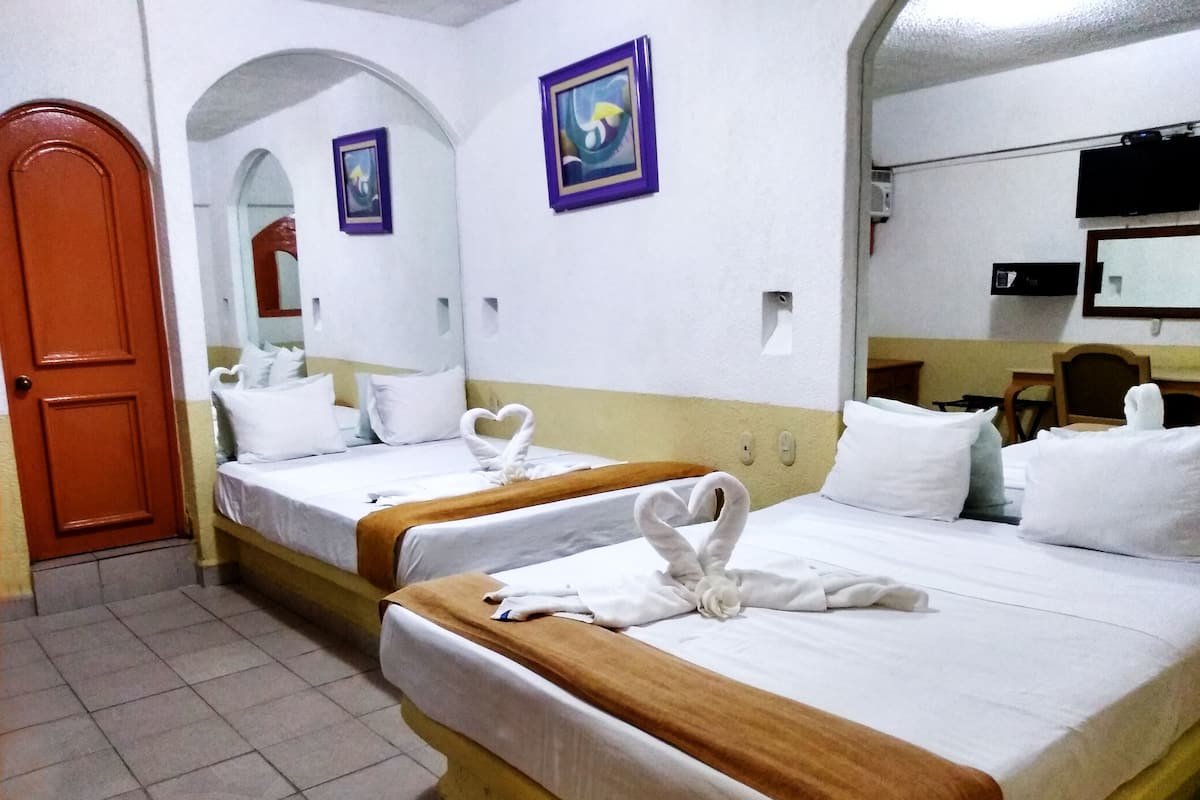 A hotel room with two beds, each with a heart-shaped towel and white bedding. There is a wooden door, a mirror, and a television on the wall.