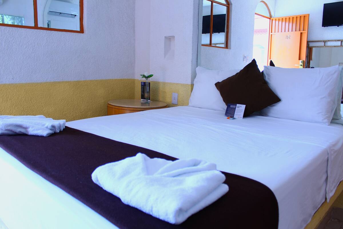 A hotel room with a neatly made bed, a bedside table with a glass of water, a small plant, and a TV mounted on the wall.
