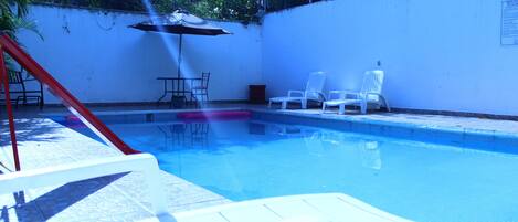 Una piscina con sillones de descanso y una mesa, rodeada por un muro blanco y vegetación.