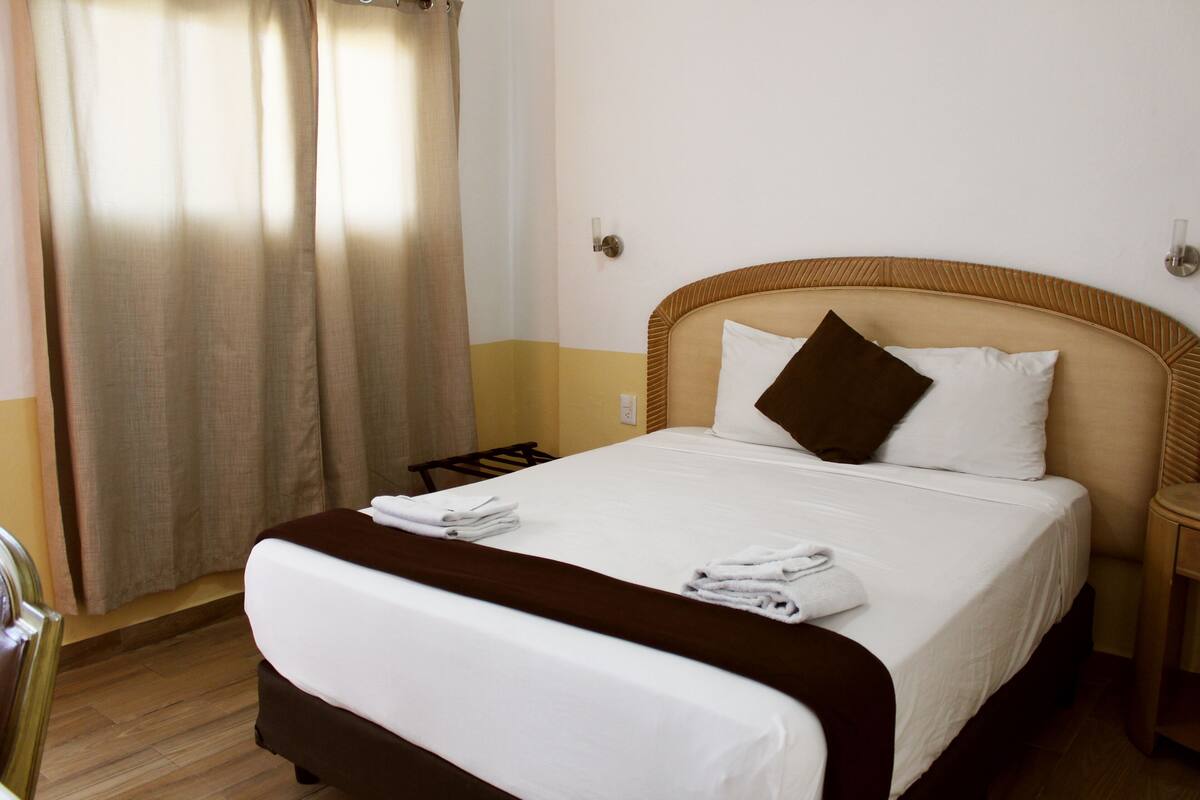 A hotel room with a bed, a chair, a nightstand, and a window with curtains.
