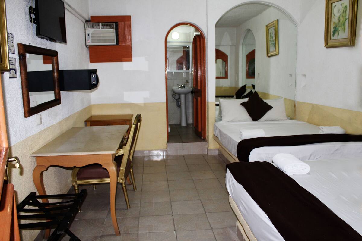 A hotel room with a bed, a desk, a chair, a mirror, and a bathroom visible through an archway.