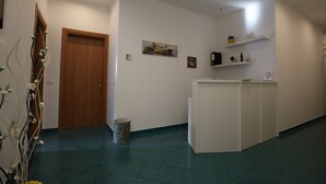 Un pasillo con una puerta de madera, un cubo de basura, un mostrador de recepción blanco y un estante de pared con diversos objetos.
