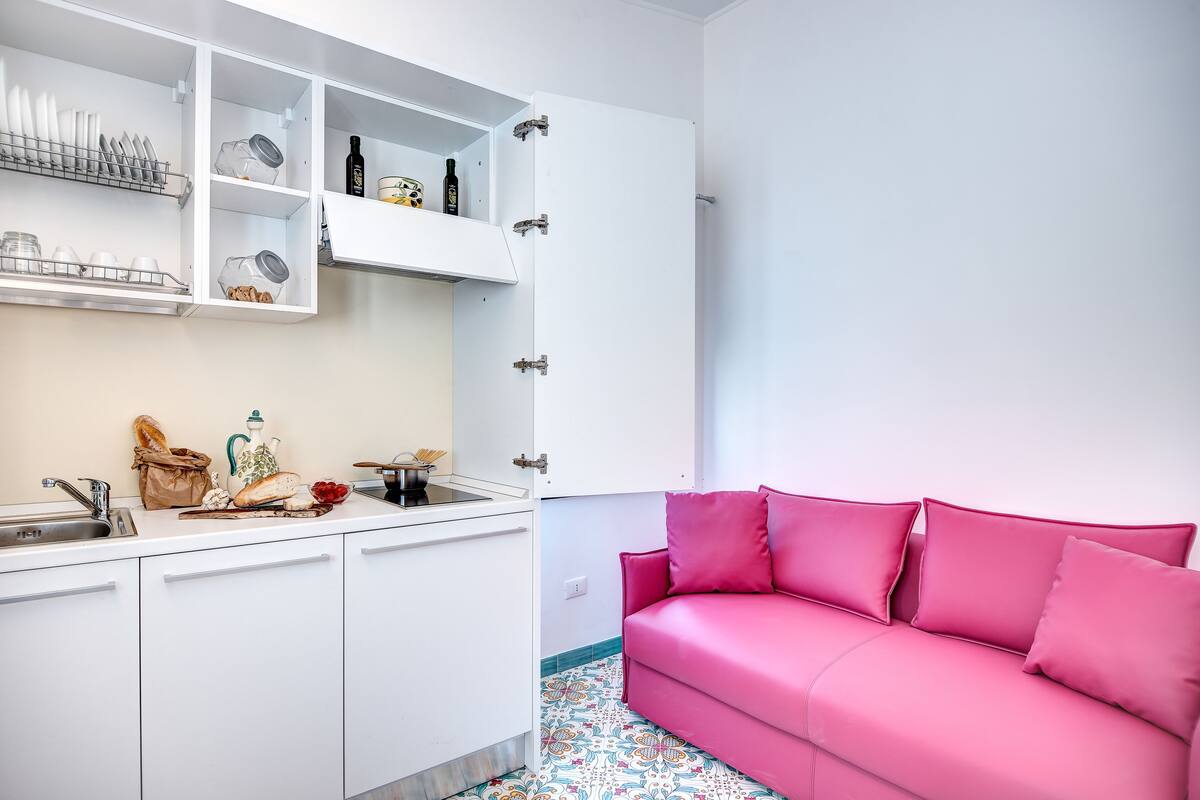 Een kleine keuken met witte kasten, een spoelbak en een roze bank.