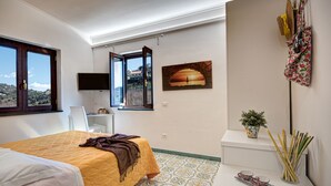 Un dormitorio con cama, escritorio, televisor, ventana con puerta abierta, cuadro, planta y un perchero en la pared.