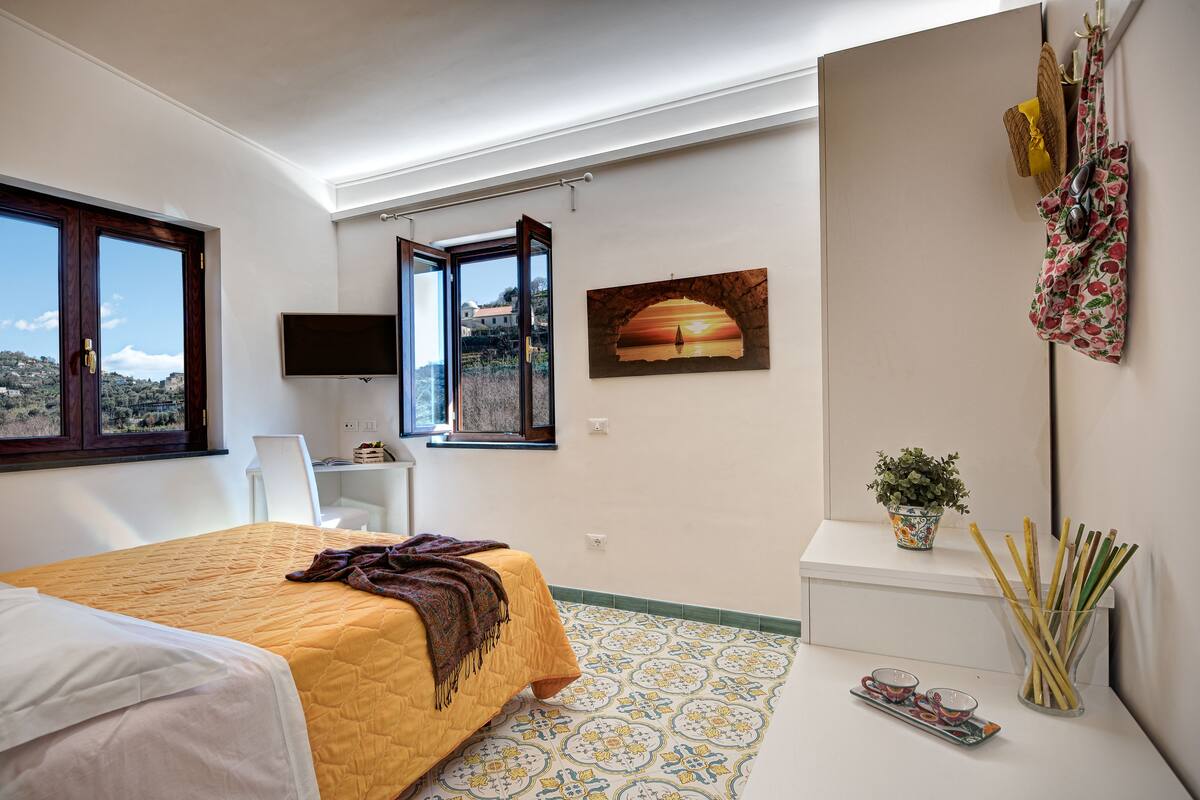 Een slaapkamer met een bed, bureau, televisie, raam met open deur, schilderij, plant en een tasrek aan de muur.