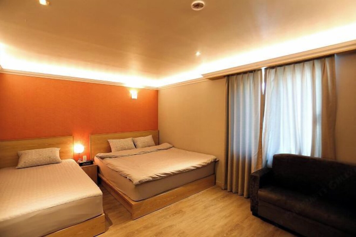 Ein Hotelzimmer mit zwei Betten, einem Schlafsofa, einem Nachttisch und einem Fenster mit Vorhängen.