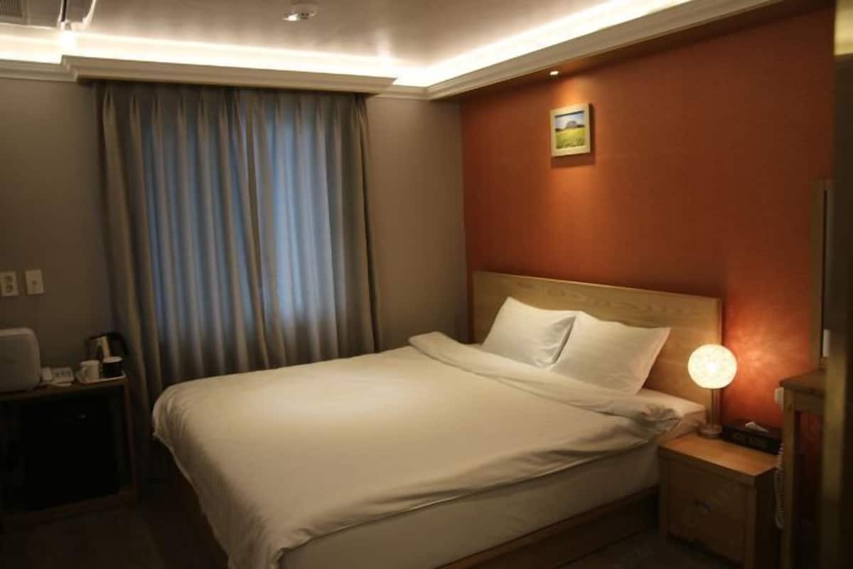 Ein Hotelzimmer mit Bett, Nachttisch mit Lampe, Schreibtisch mit Stuhl und einem Fenster mit Vorhängen.