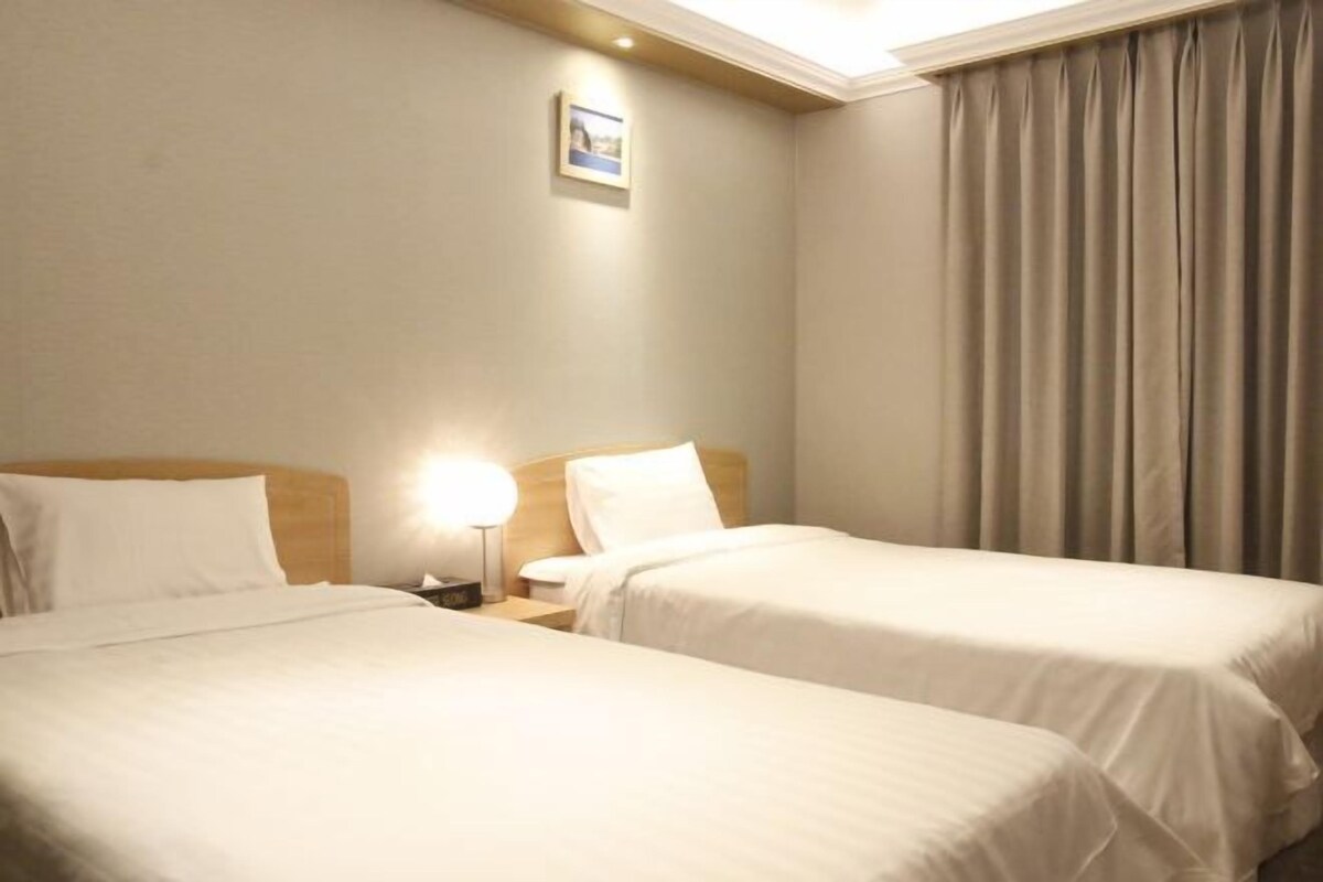 A hotel room with two beds, a nightstand with a lamp, a framed picture on the wall, and a large window with curtains.