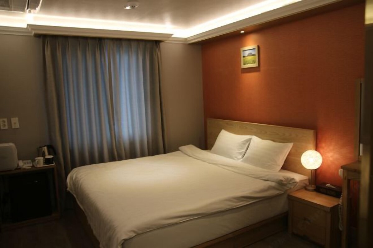 Ein Hotelzimmer mit Bett, Nachttisch mit Lampe, Schreibtisch mit Stuhl und einem Fenster mit Vorhängen.