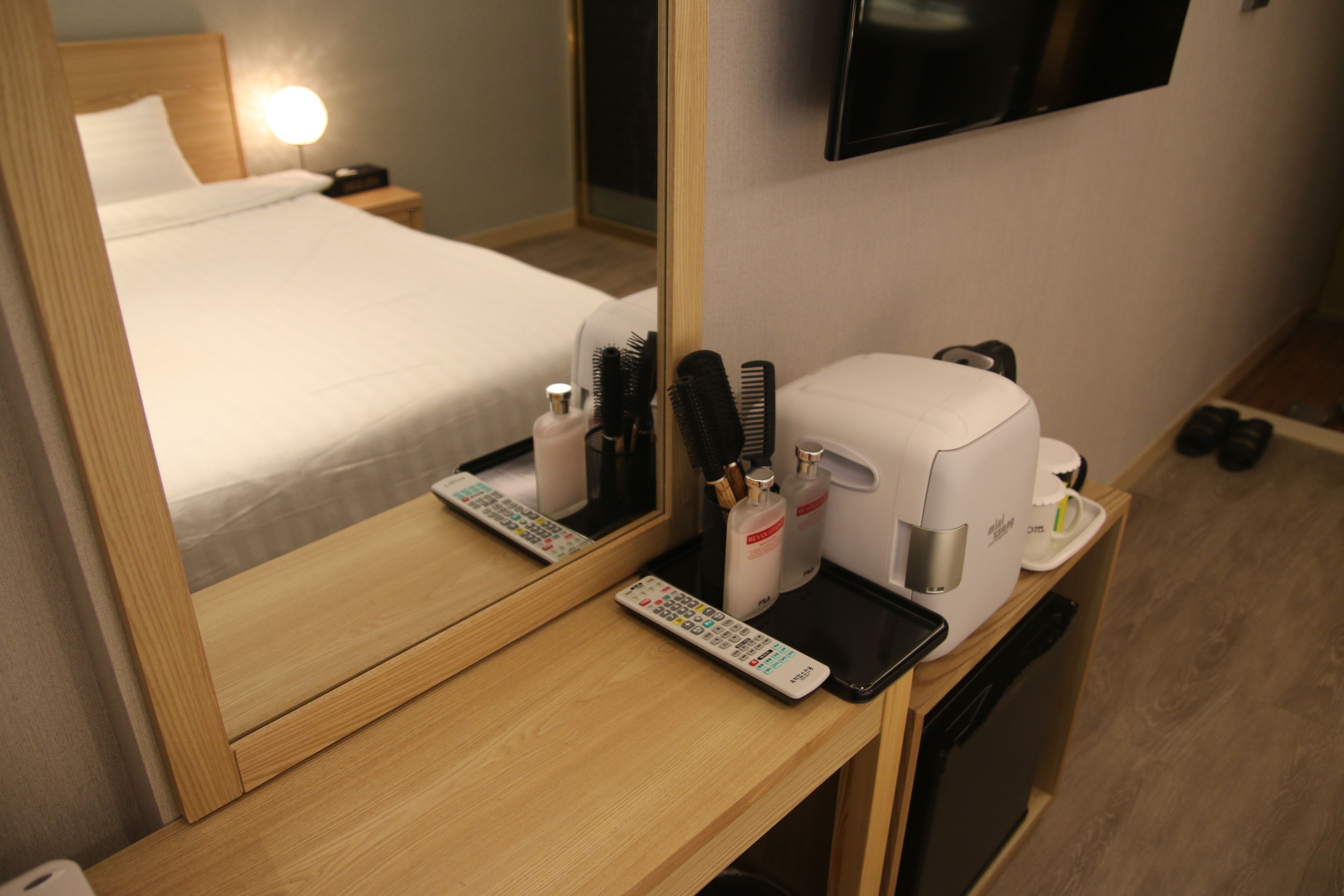 A hotel room with a wooden desk, a mirror, a bed, a TV mounted on the wall, a coffee machine, and a remote control.