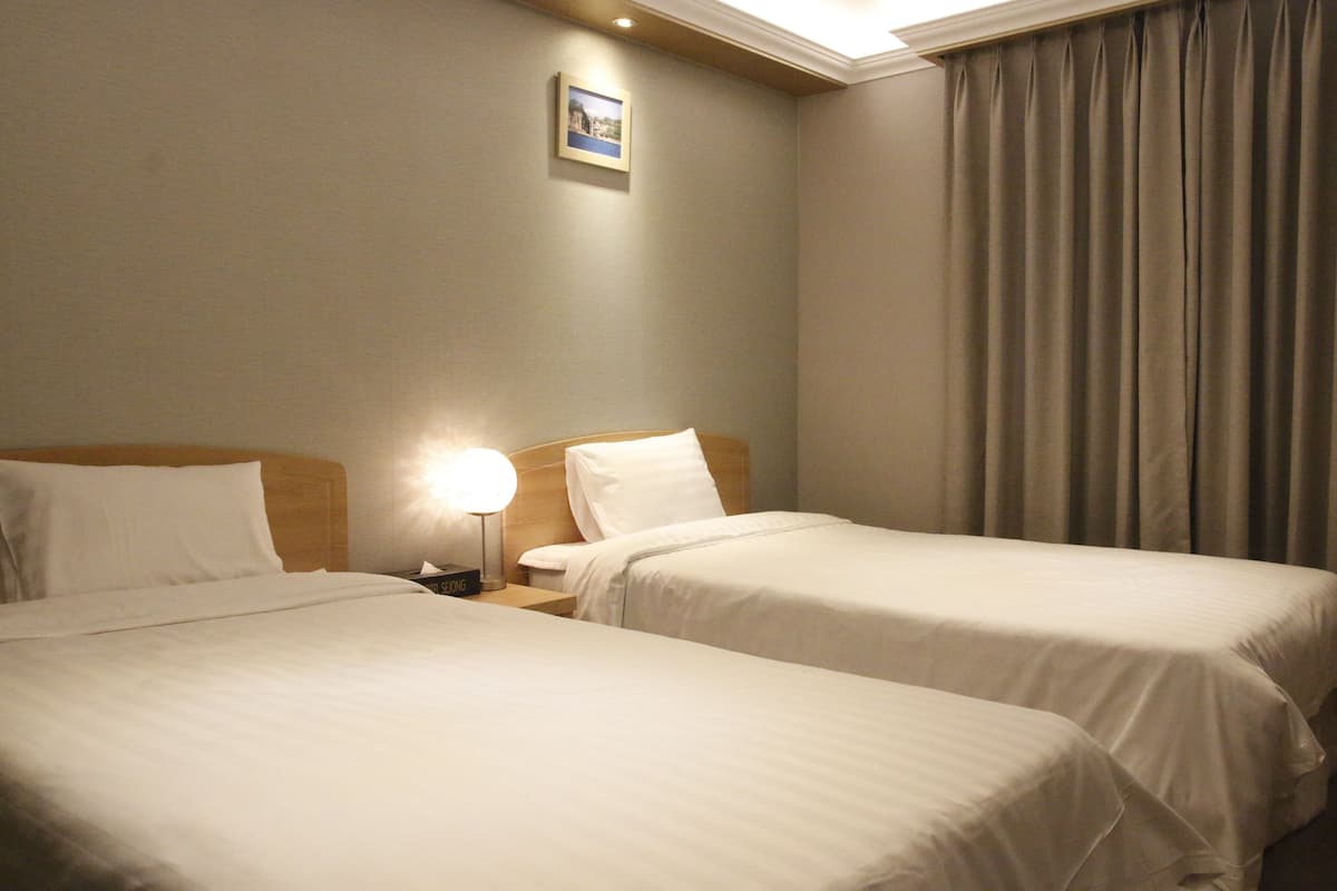 Ein Hotelzimmer mit zwei Einzelbetten, einem Nachttisch mit Lampe, einem gerahmten Bild an der Wand und einem Fenster mit Vorhängen.