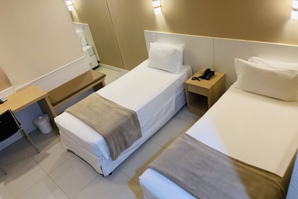 A hotel room with two beds, a bedside table with a phone, a small table with a lamp, and a trash bin.