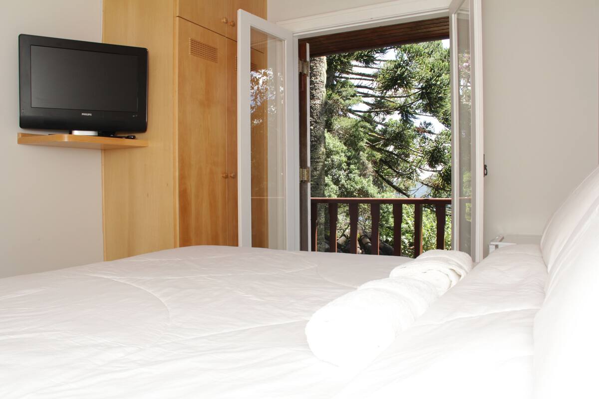 Un dormitorio con cama, televisor montado en la pared y balcón con vistas a árboles.