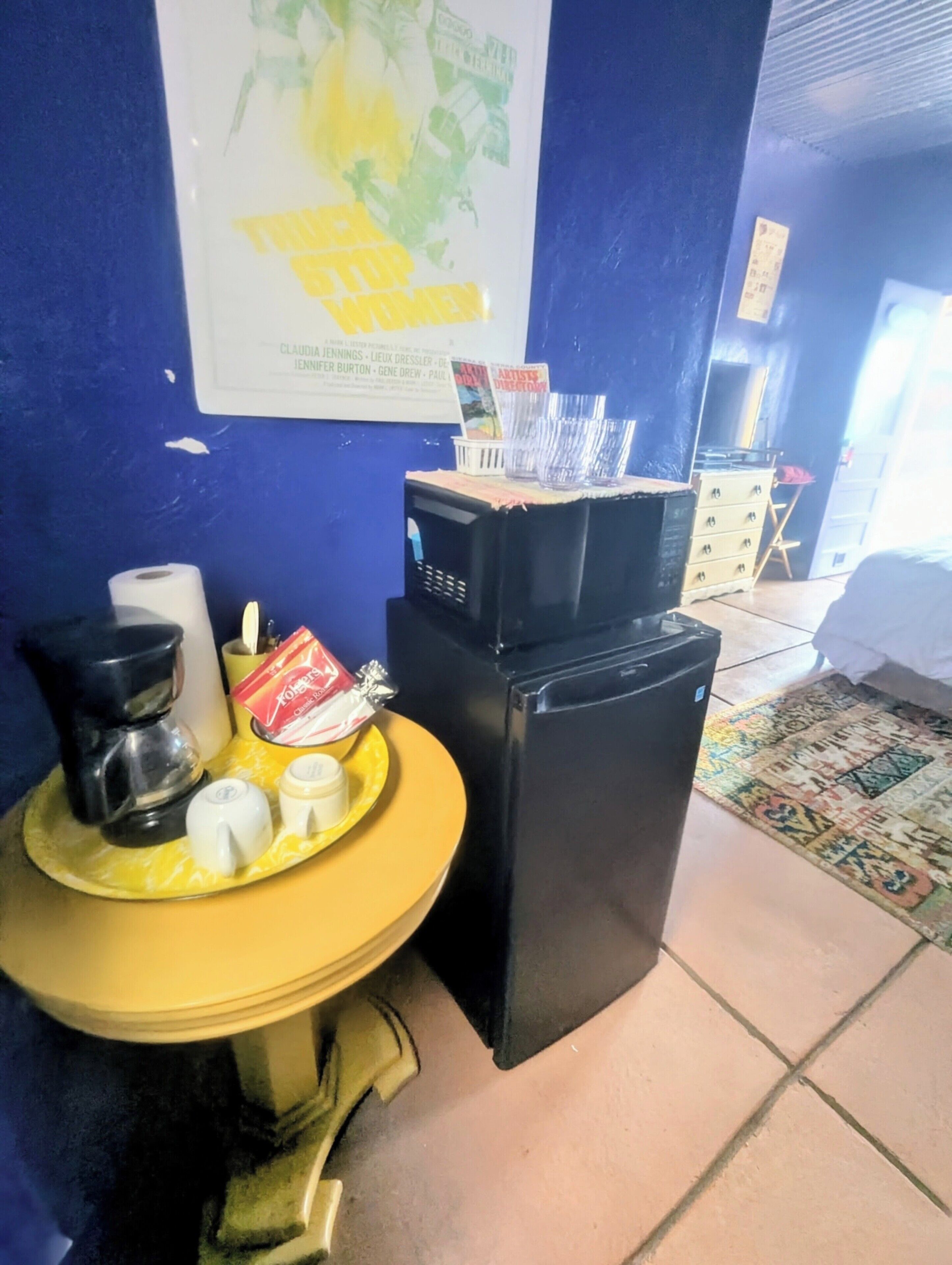 Una kitchenette compacta con cafetera, microondas, refrigerador pequeño y una mesa amarilla con aderezos.