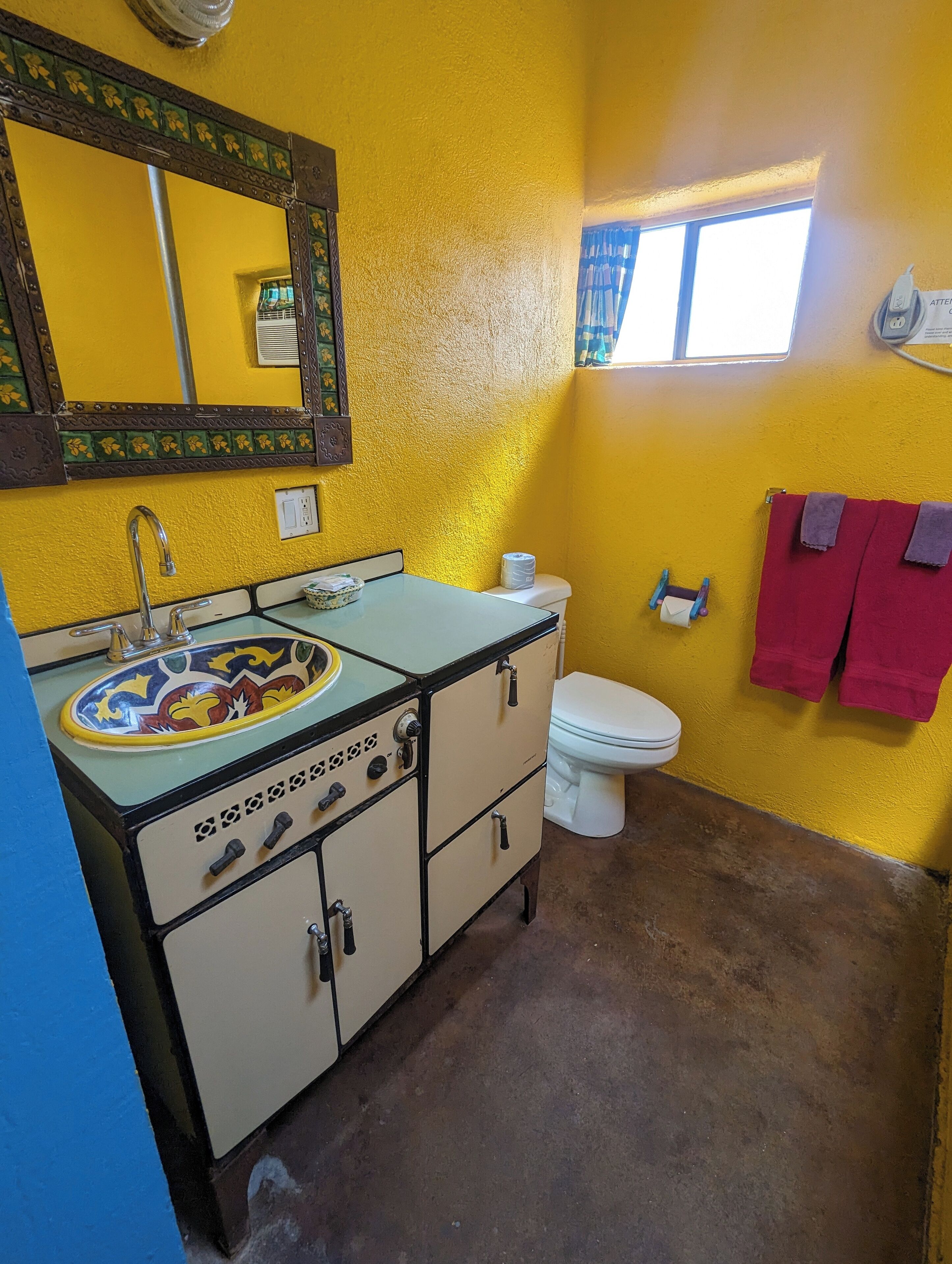 A bathroom with a yellow wall, a sink with a decorative basin, a toilet, and a window with curtains.