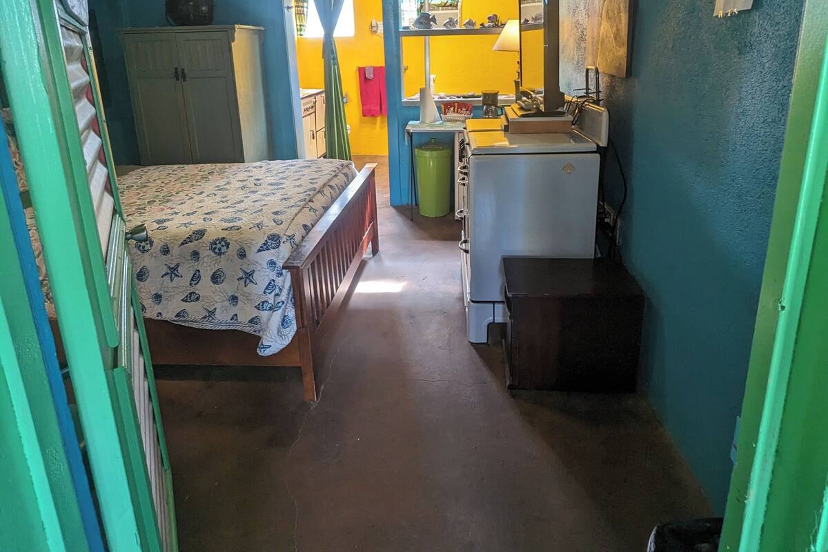 A compact room with a bed, a small desk, a mirror, and a kitchenette.