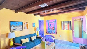 A living room with a wooden ceiling, a blue sofa, a purple armchair, a coffee table, and a colorful abstract painting on the wall.