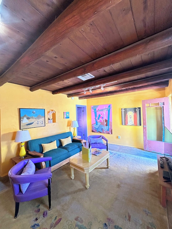 Een woonkamer met een houten plafond, een blauwe bank, een paarse fauteuil, een salontafel en een kleurrijk abstract schilderij aan de muur.