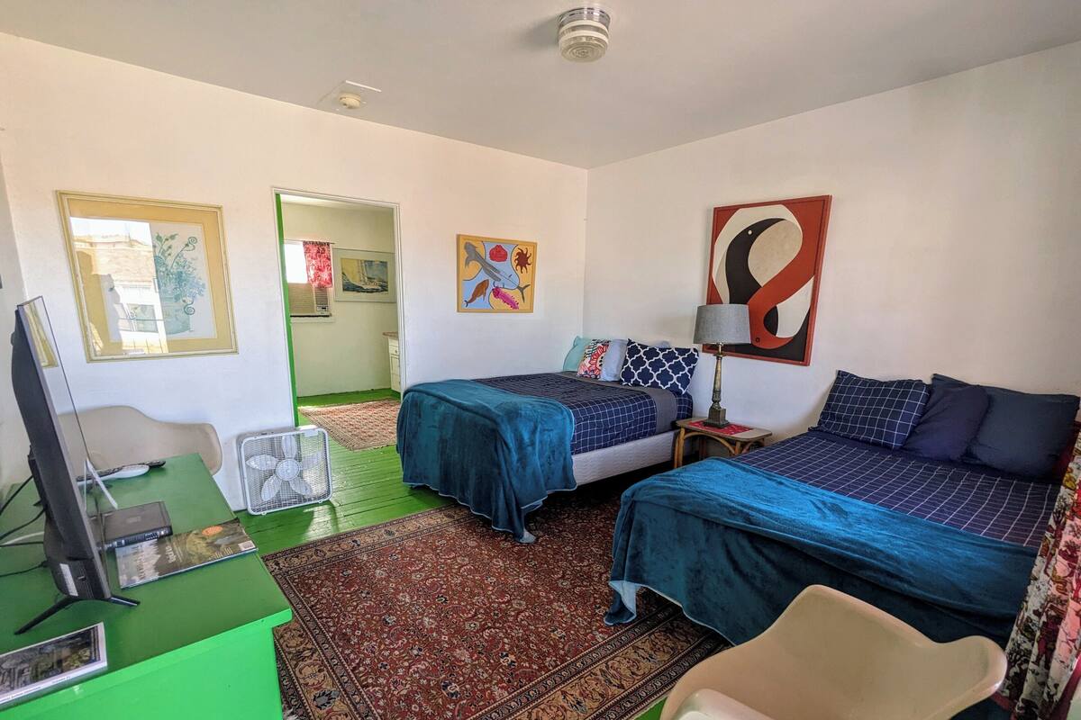 A hotel room with two beds, a desk with a computer, a chair, a fan, and a rug.