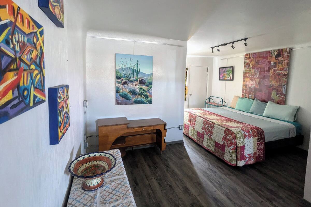 A bedroom with a bed, a desk, and a chair. There are paintings on the walls and a window with curtains.