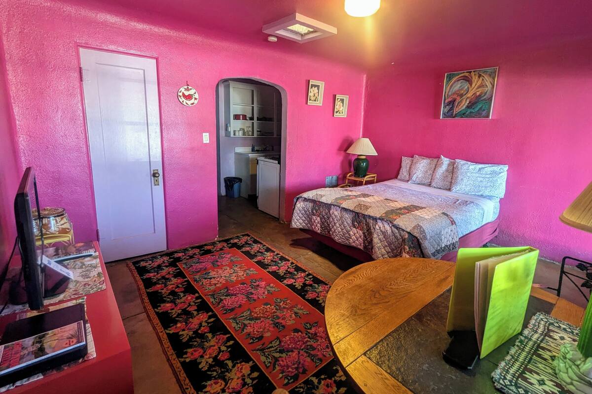 A pink-themed room with a bed, a desk, a chair, a lamp, and a small kitchen area.
