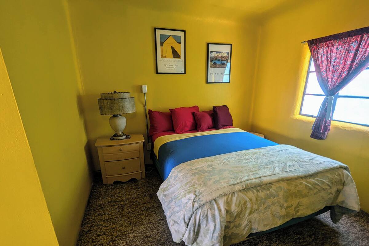 A bedroom with a bed, a nightstand, a lamp, two framed pictures on the wall, and a window with a curtain.