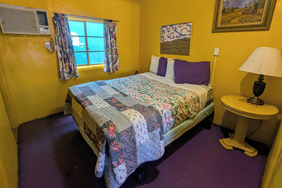 A room with a bed, a window with curtains, a painting on the wall, and a lamp on a side table.