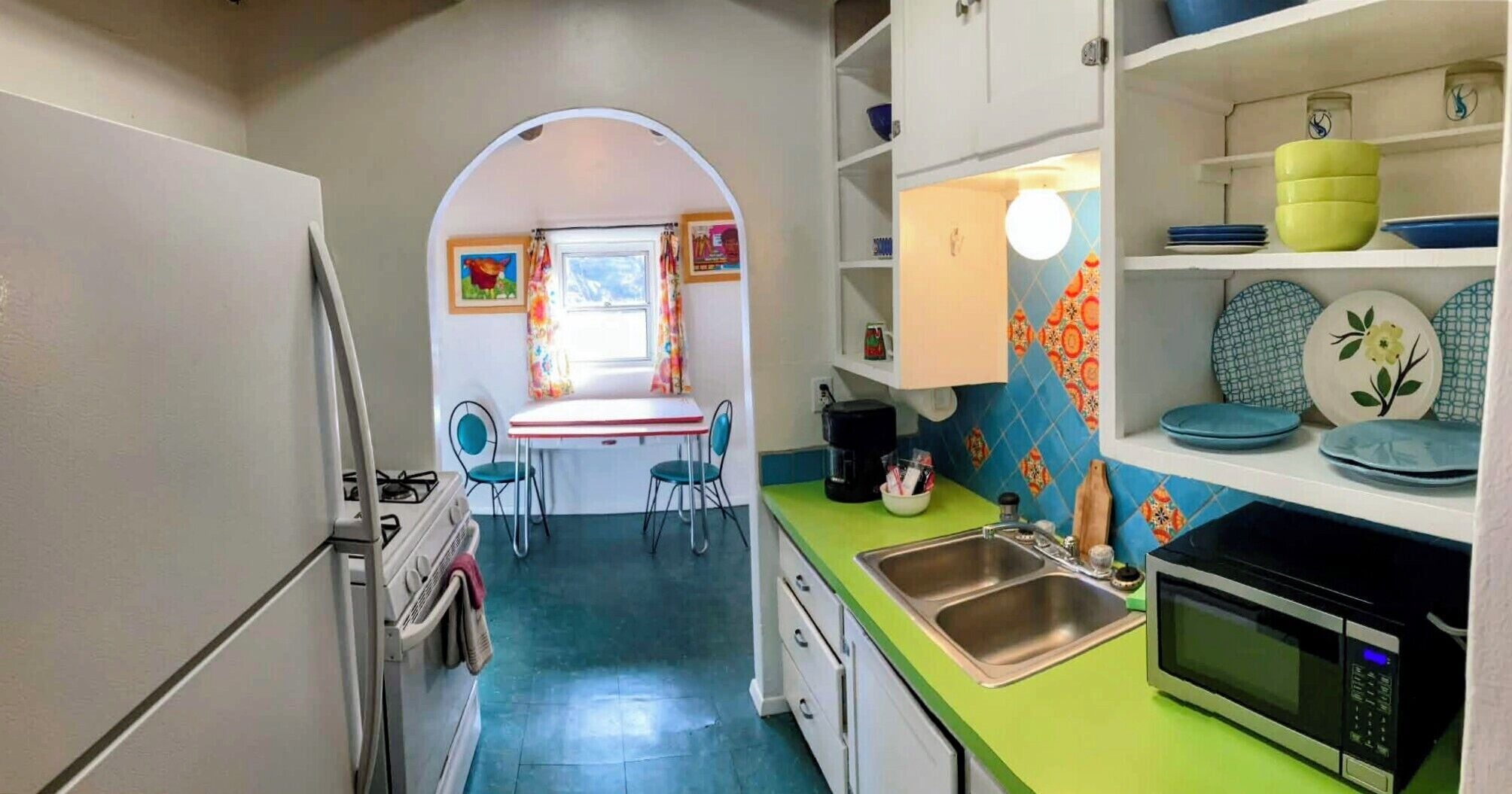 A compact kitchen with a gas stove, a microwave, and a sink. There is a dining area with a table and chairs, and a colorful wall decoration.