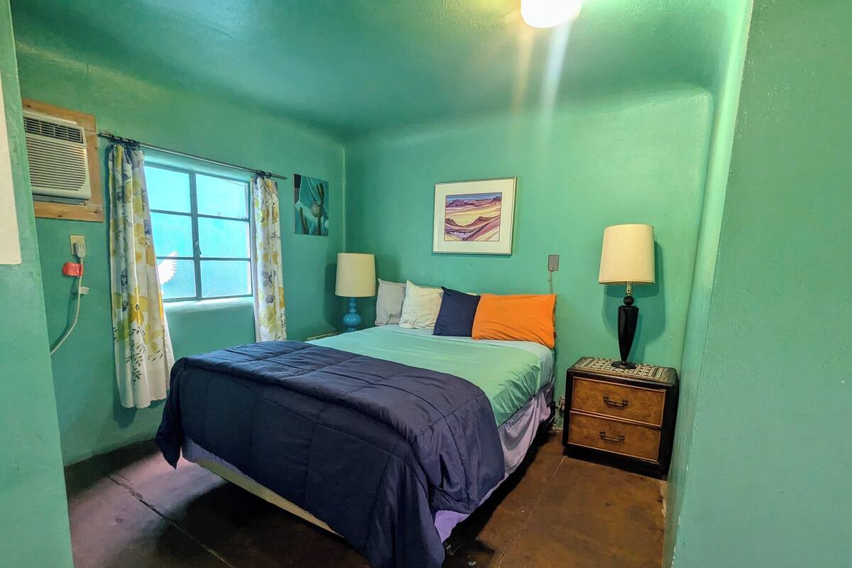 A bedroom with a bed, two bedside tables, a window with curtains, a wall-mounted air conditioner, and a wall painting.