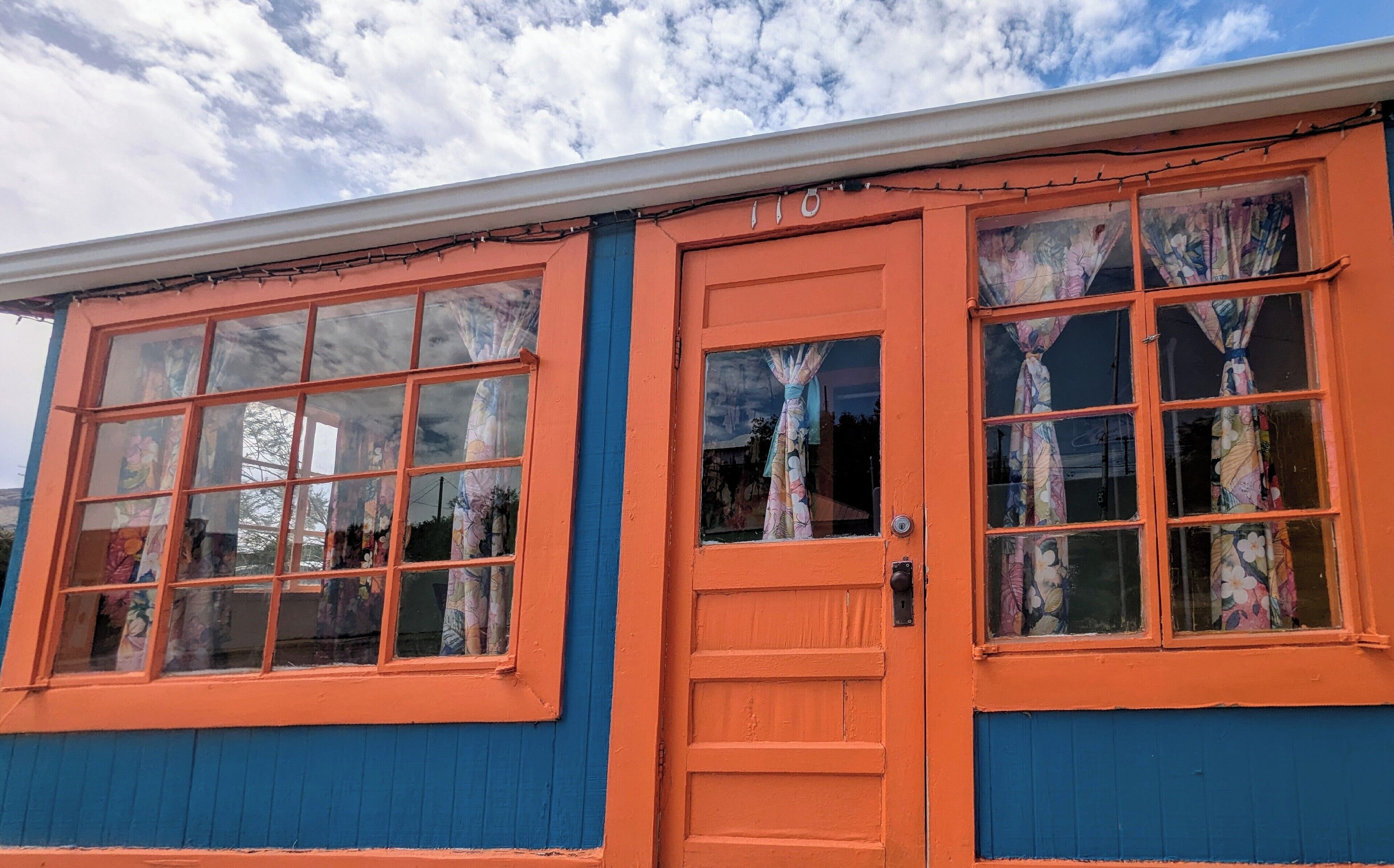 A building with a blue lower half and an orange upper half, featuring large windows with floral curtains.