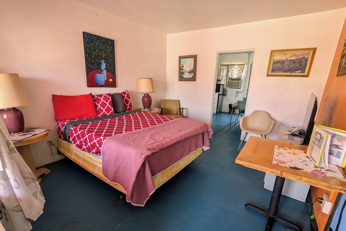 A hotel room with a bed, two bedside tables, a desk, and a chair. There are paintings on the walls and a window with curtains.