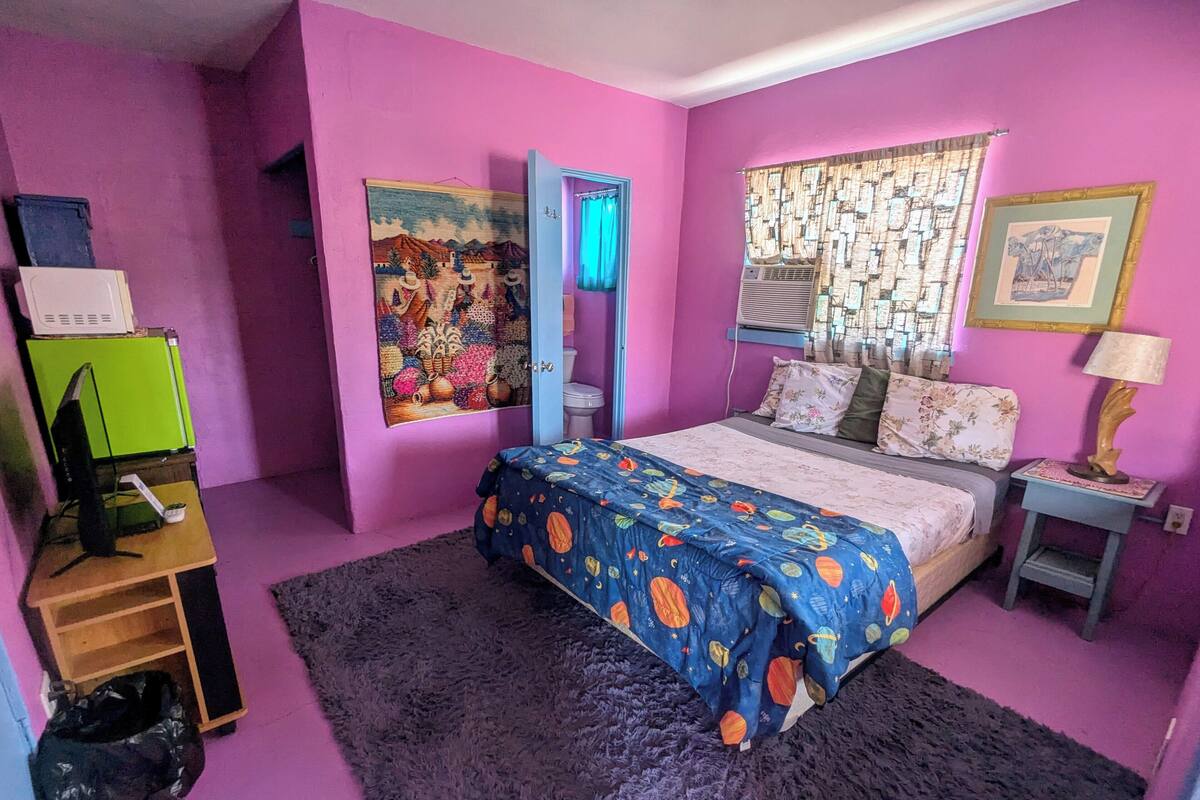 A bedroom with a bed, a desk, a chair, a window with curtains, and a painting on the wall.