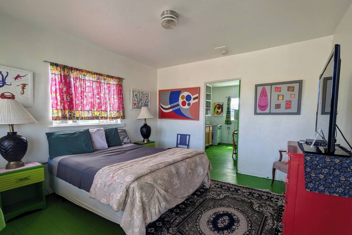 A bedroom with a bed, green walls, a window with colorful curtains, a desk with a computer, and artwork on the walls.