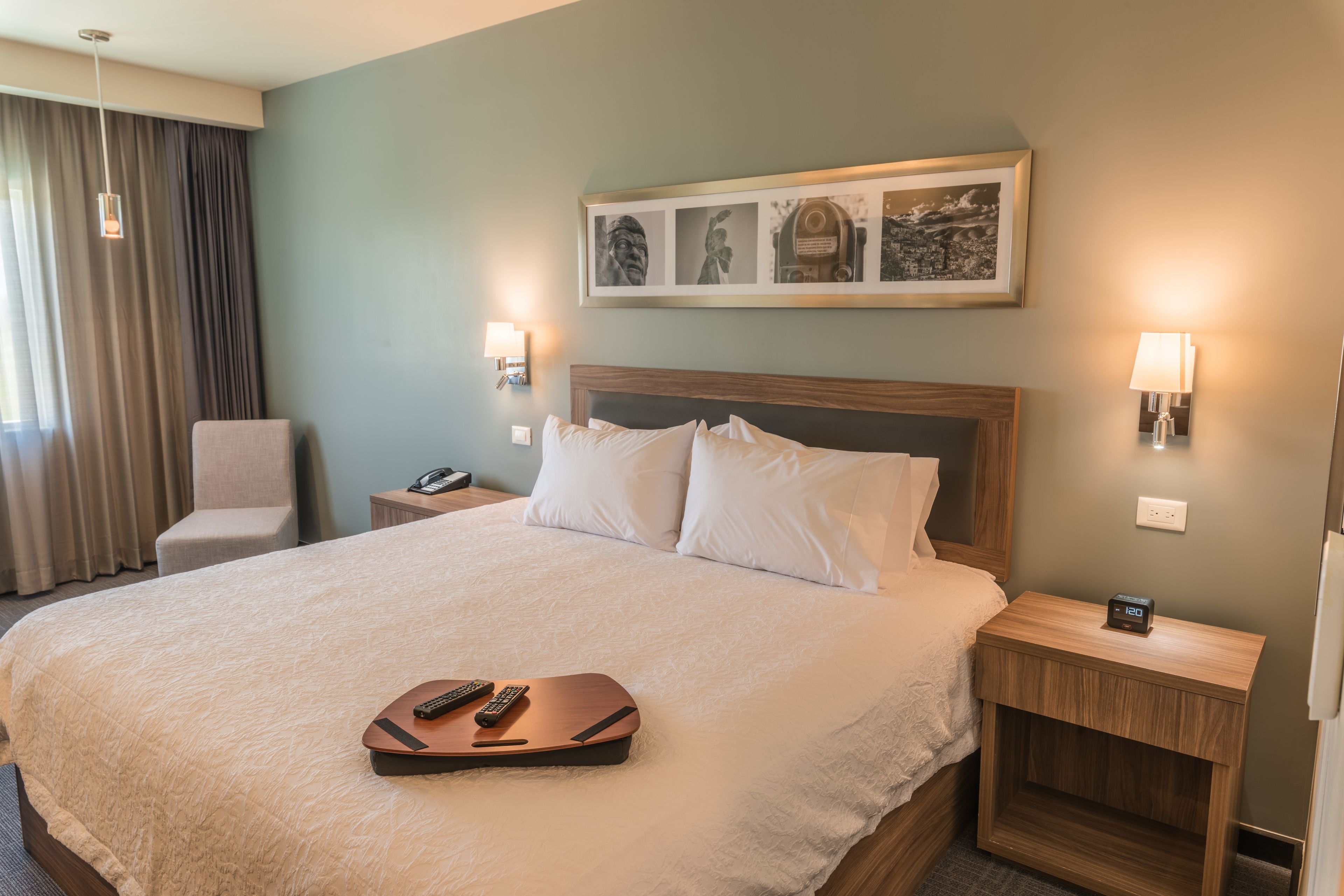 A hotel room with a large bed, a chair, a nightstand with a phone, a lamp, and a framed picture on the wall.