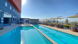 A swimming pool with clear blue water, surrounded by lounge chairs and umbrellas, and a modern building with a red facade in the background. - Hampton Inn & Suites by Hilton Salamanca Bajio (Salamanca)