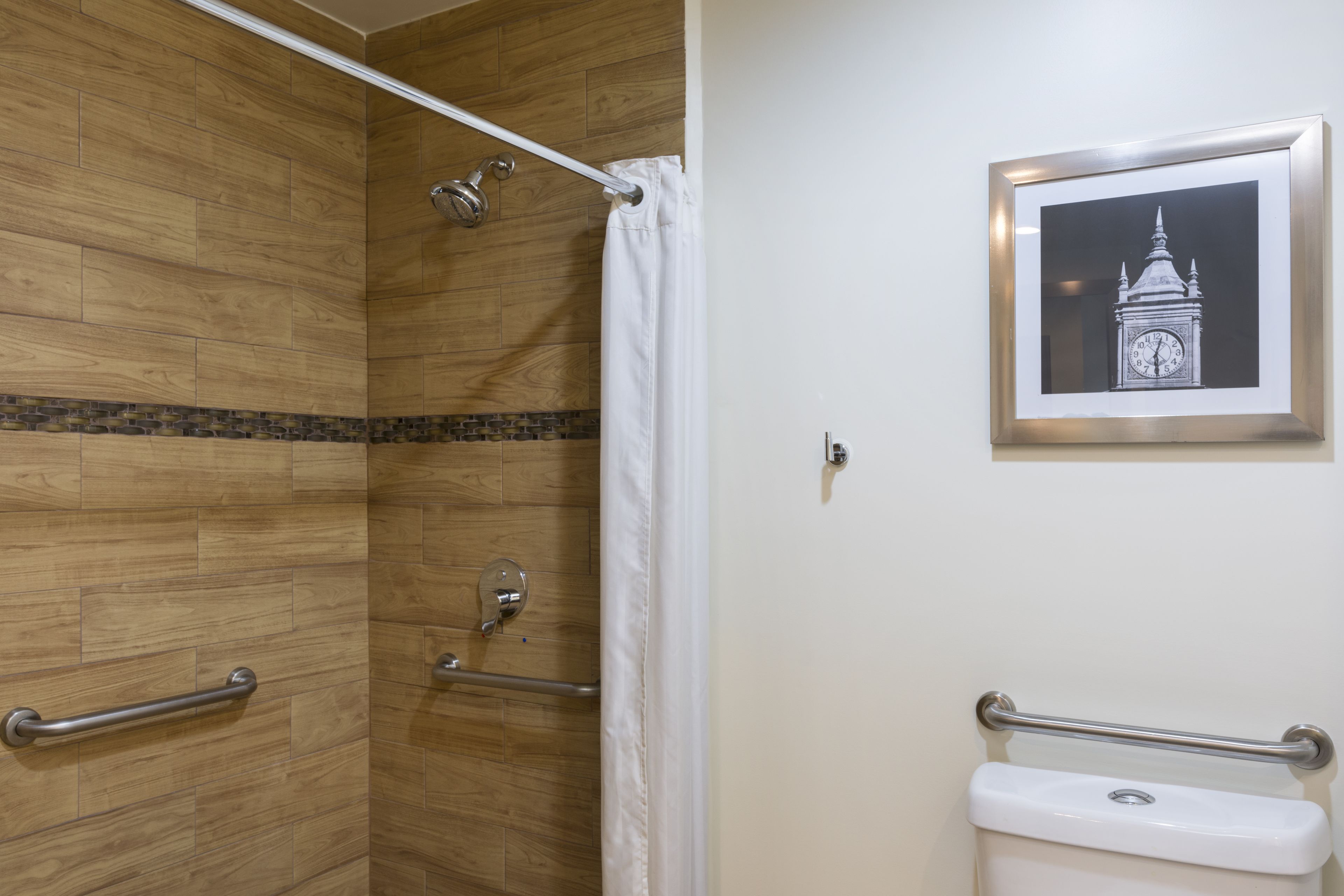 Un baño con suelo de madera, ducha con puerta de cristal, inodoro con barra de apoyo y un cuadro con una torre del reloj en la pared.