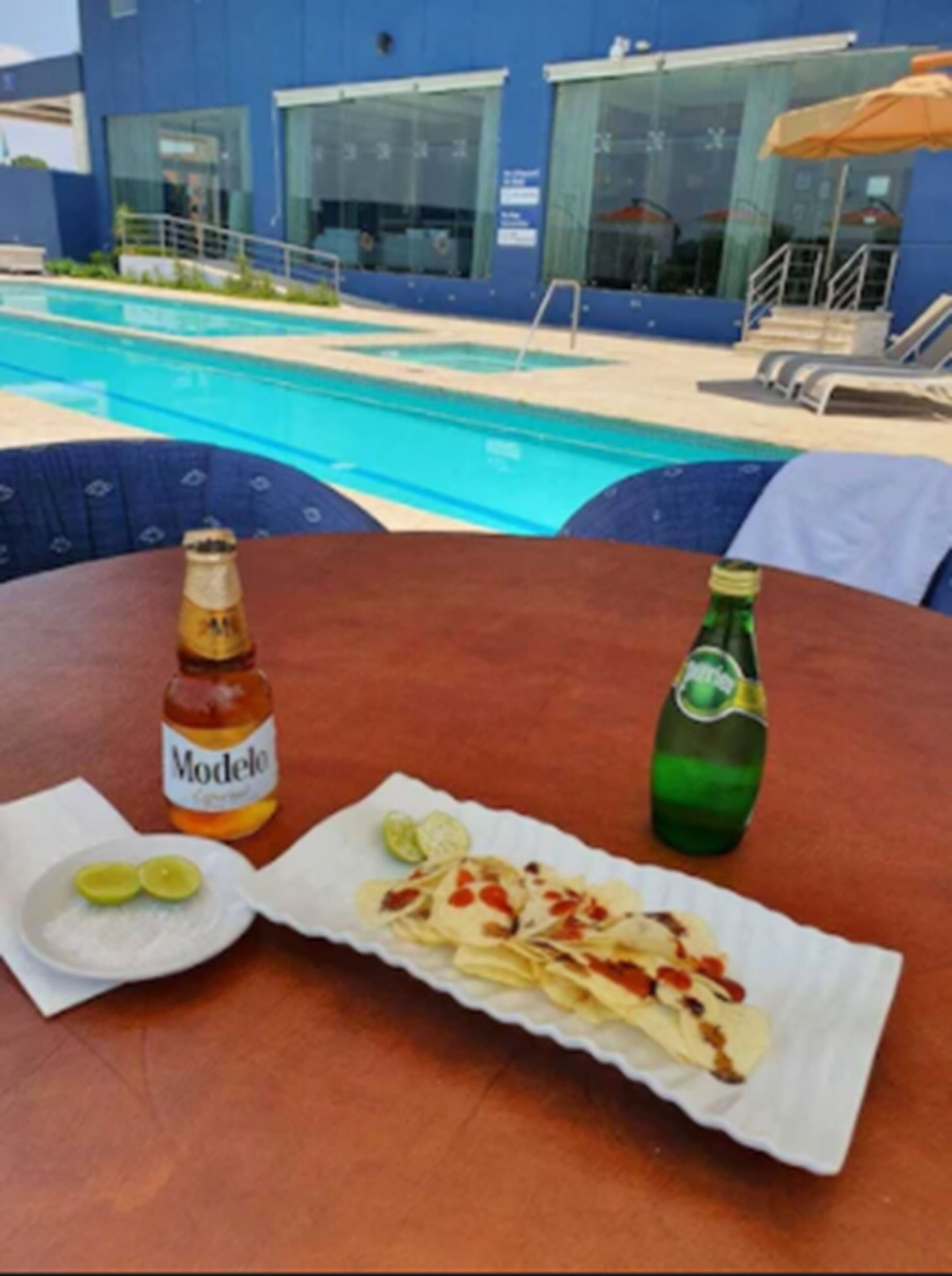 A table with a bottle of Modelo beer, a plate with lime slices, and a wrapped food item, overlooking a pool area with lounge chairs and umbrellas.