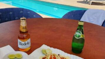 A table with a bottle of Modelo beer, a plate with lime slices, and a wrapped food item, overlooking a pool area with lounge chairs and umbrellas.