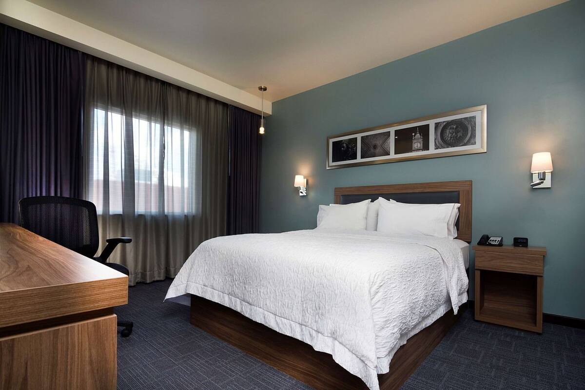 A hotel room with a bed, a desk, a chair, a nightstand, and a wall-mounted lamp.