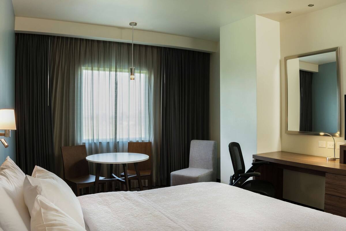 A hotel room with a bed, a desk with a TV, a chair, a small table, and a window with curtains.
