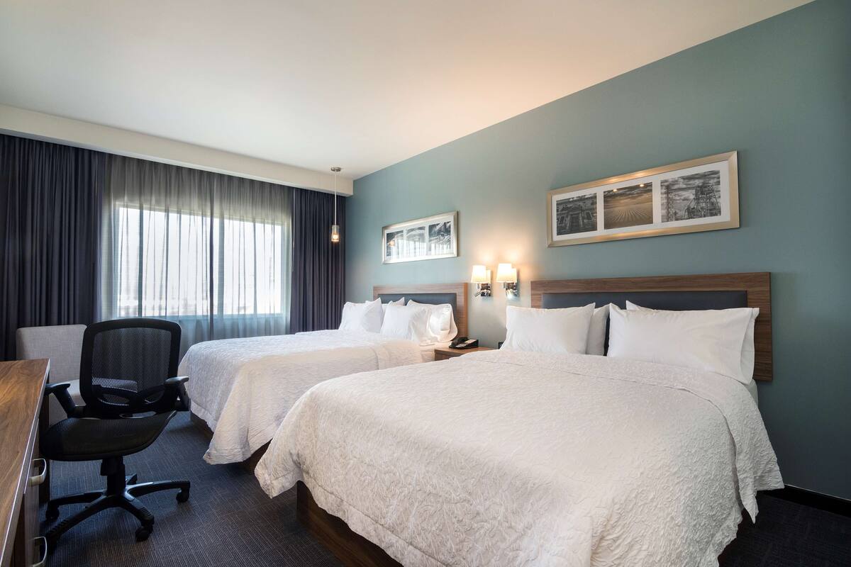 A hotel room with two beds, a desk, a chair, and a large window with curtains.