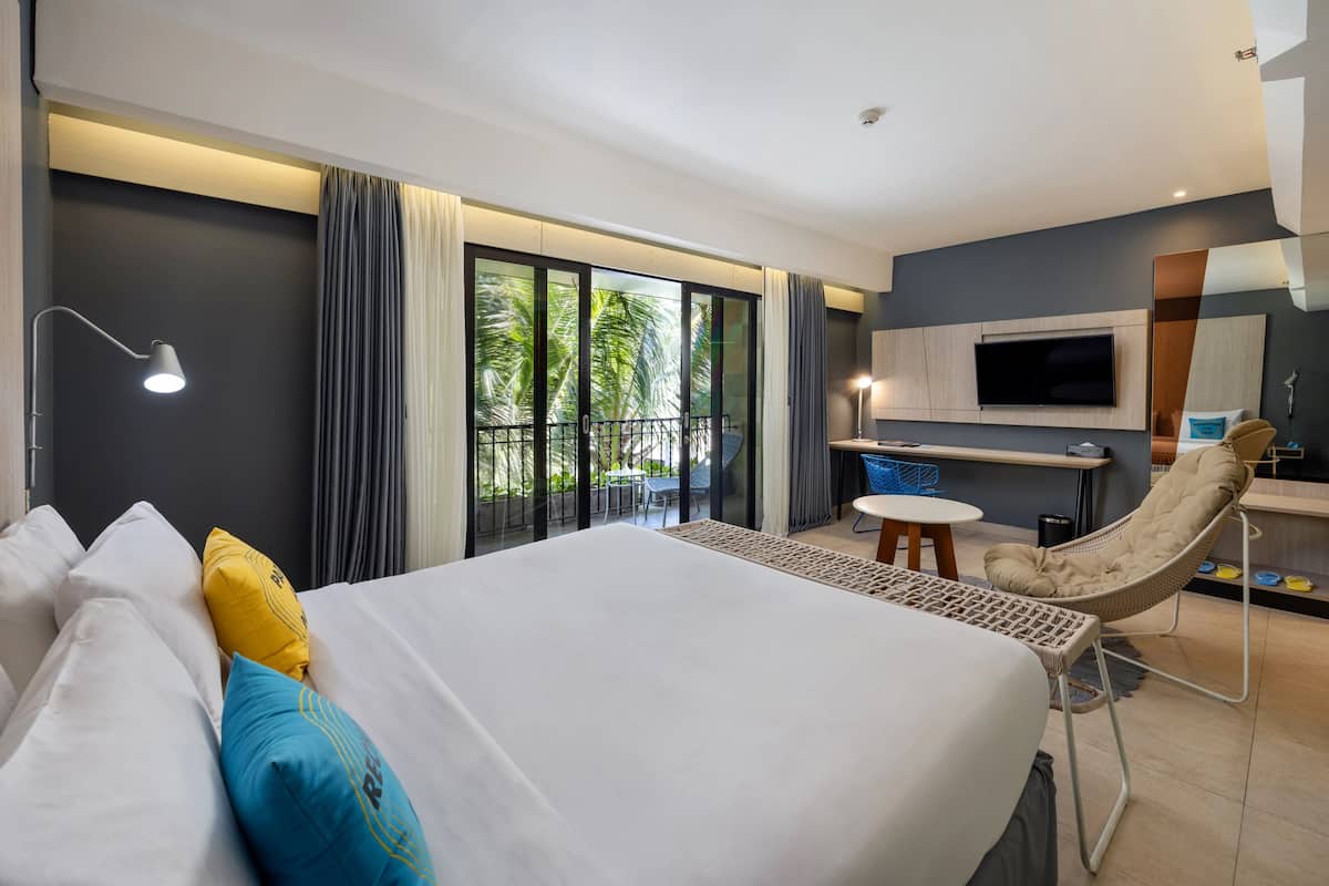 A modern hotel room with a large bed, a desk with a chair, a TV, and a balcony with a view of palm trees.