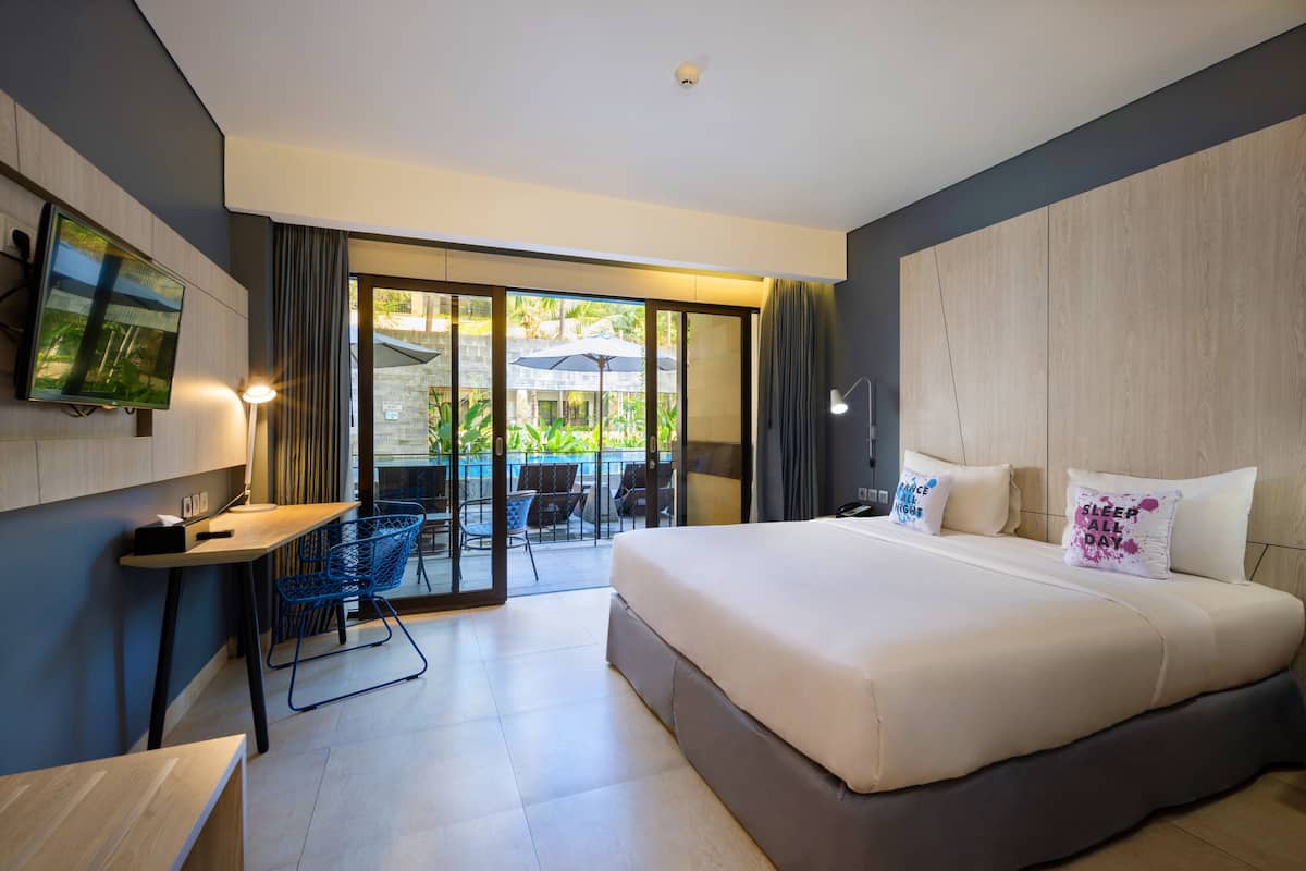 A modern hotel room with a large bed, a desk, a TV, and a balcony with outdoor seating.