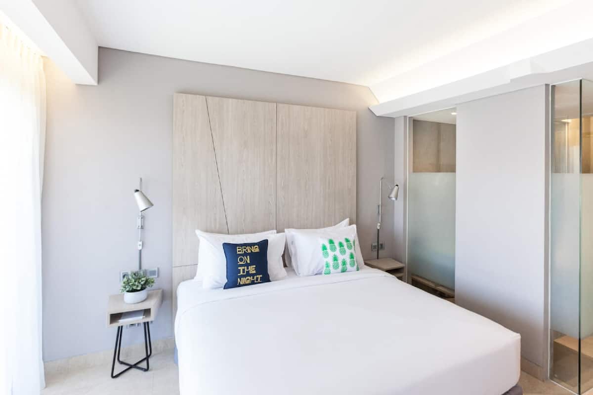 A modern hotel room with a large bed, a nightstand with a lamp, and a glass shower enclosure.