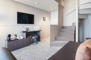 A modern hotel room with a flat-screen TV, a small kitchenette, a seating area with a sofa and a chair, and a staircase.