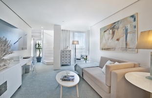 A modern hotel room with a minimalist design, featuring a sofa, a coffee table, a desk, and a large abstract painting on the wall.