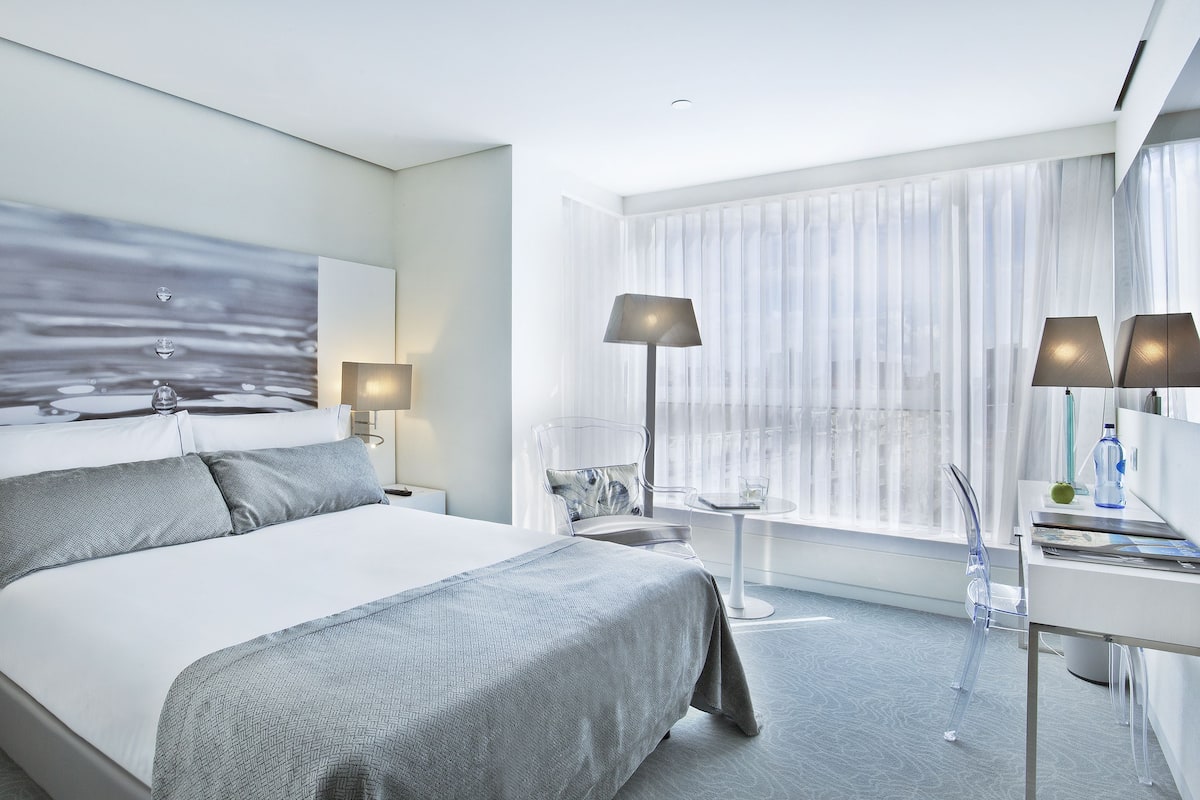Een moderne hotelkamer met een groot bed, een bureau en een stoel. Er is een raam met dunne gordijnen en uitzicht op de stad.