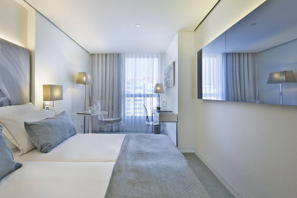 Een moderne hotelkamer met een groot bed, een bureau en een stoel. Er staan twee lampen op het bureau en een spiegel aan de muur.