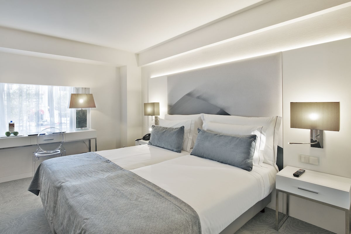 Een moderne hotelkamer met een groot bed, nachtkastjes, een bureau en een raam met dunne gordijnen.